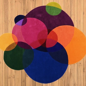May include: A vibrant, abstract rug with a design of overlapping circles in various colours including orange, pink, purple, blue, green, and yellow. The rug has a soft texture and is designed to add a splash of colour to a room. It is placed on a wooden floor.