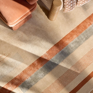 Pode incluir: Tapete bege e castanho claro com riscas ferrugem e cinzentas. O tapete tem um padrão geométrico e é combinado com uma poltrona cor de pêssego e um objeto cilíndrico de madeira. Um cobertor estampado também é visível.