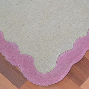 Könnte beinhalten: Nahaufnahme eines hellgrünen Teppichs mit einem wellenförmigen, rosa Rand. Der Teppich liegt auf einem Holzboden mit diagonalem Muster. Das Design ist schlicht und elegant.
