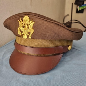 Könnte beinhalten: Ein brauner Militärhut mit goldenem Emblem und braunem Lederschirm. Der Hut hat ein goldenes Band und zwei goldene Knöpfe. Der Hut ist ein klassischer Militärstil.