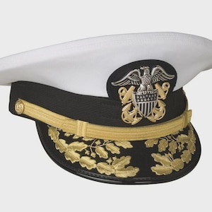 Könnte beinhalten: Eine weiß-schwarze Militärmütze mit goldenem Kinnriemen und einem silber-goldenen Adleremblem auf der Vorderseite. Der schwarze Schirm ist entlang der Vorderkante mit goldfarbenen Stickereien von Eichenblättern und Eicheln verziert.