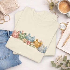 May include: Two cream-colored t-shirts with a floral design featuring colorful hen-shaped containers. The hens are in shades of pink, green, blue, and yellow, with a floral arrangement. The shirts have the text "Sass'n Sunshine" on a sticker.