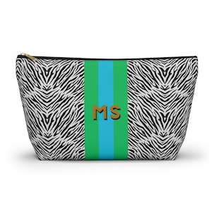 May include: A zippered pouch with a zebra print pattern. The center features a green and blue stripe with the letters "MS" in a brown color. The zipper is gold.