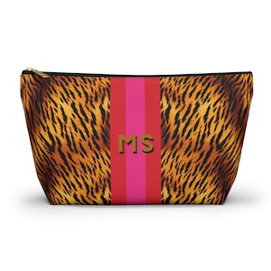 May include: A zippered cosmetic bag with a tiger stripe pattern in shades of brown and orange. A vertical stripe of pink and red runs down the center, with the gold letters "MS" in the middle. The bag has a black zipper.