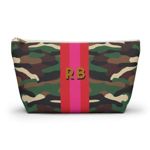 May include: A camouflage print zippered pouch with a vertical stripe in pink and red. The stripe features the gold letters "RB". The pouch has a gold zipper and is designed to hold various items.
