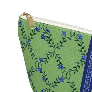 May include: A green cosmetic bag with a floral pattern of blue flowers and green vines. The bag has a gold zipper and a navy blue border with a Greek key design. The bag is made of a textured fabric.