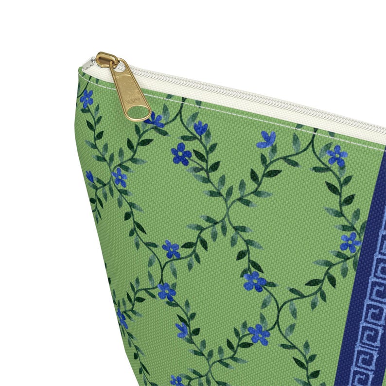 May include: A green cosmetic bag with a floral pattern of blue flowers and green vines. The bag has a gold zipper and a navy blue border with a Greek key design. The bag is made of a textured fabric.