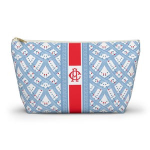 May include: A zippered pouch with a light blue and white lattice pattern, accented with red dots. A central red stripe features a monogram. The pouch has a gold-colored zipper.