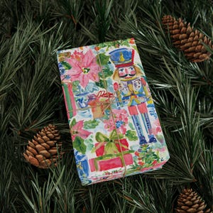May include: A rectangular gift box wrapped in white paper featuring a colorful watercolor print of a nutcracker, pink poinsettia flowers, and red gift boxes, tied with thin brown twine across the center of the package.
