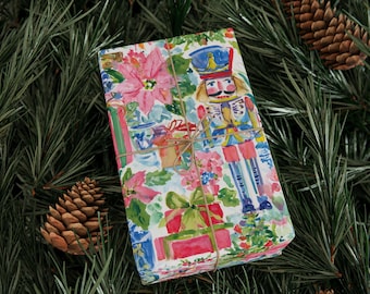 May include: A rectangular gift box wrapped in white paper featuring a colorful watercolor print of a nutcracker, pink poinsettia flowers, and red gift boxes, tied with thin brown twine across the center of the package.