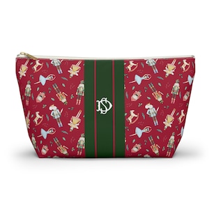 Könnte beinhalten: Eine rote Kosmetiktasche mit goldenem Reißverschluss und einem festlichen Muster aus Nussknackern, Schaukelpferden und Ballettschuhen. Ein grüner Streifen mit weißem Monogramm verläuft in der Mitte.
