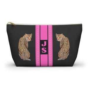 Könnte beinhalten: Schwarze Reißverschlusstasche mit zwei Leoparden-Illustrationen und einem rosa vertikalen Streifen mit den Buchstaben "JS". Der Streifen ist von einem griechischen Schlüsseldesign umrandet. Die Tasche hat einen goldenen Reißverschluss.