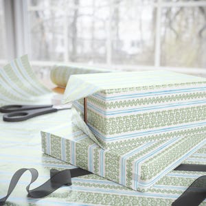 May include: Two rectangular boxes wrapped in white paper featuring a repeating pattern of vertical light blue stripes and intricate green floral bands. A roll of matching wrapping paper and black scissors are visible nearby on the surface.