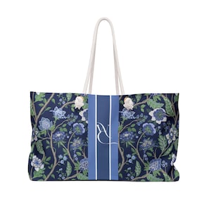 Op de afbeelding: Grote draagtas met een bloemenpatroon in blauw, groen en wit op een marineblauwe achtergrond. De tas heeft een verticale streep met initialen en touwhandvatten.