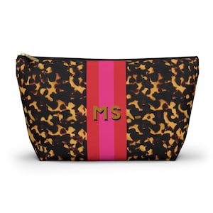 May include: A zippered cosmetic bag with a tortoiseshell pattern and a vertical stripe in red and pink. The bag features gold-colored initials "MS" in the center of the stripe. The bag is designed to hold makeup and other beauty products.
