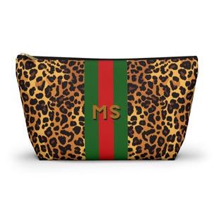 Peut inclure: Une trousse de maquillage à imprimé léopard avec une bande verte et rouge au centre. Les initiales "MS" sont en lettres dorées sur la bande. La trousse a une fermeture éclair dorée.