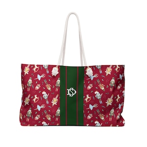 Könnte beinhalten: Große rote Tragetasche mit weißem Seilgriff. Die Tasche hat ein sich wiederholendes Muster aus Nussknackern, Schaukelpferden und anderen Illustrationen zum Thema Feiertage. Ein grüner Streifen mit weißem Monogramm verläuft durch die Mitte.