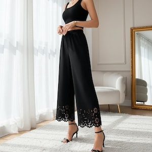 May include: Black two-piece outfit comprising a crop top and wide-leg trousers. The trousers have a floral cutout design at the hem. The outfit is paired with black high-heeled sandals. The background includes a white rug and a gold-framed mirror.