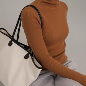 May include: A woman wearing a brown ribbed turtleneck jumper and light grey trousers. A white tote bag with black trim and handles is over the shoulder. The outfit is simple and stylish.