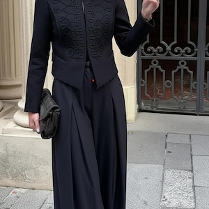 Peut inclure: Veste noire ornée de broderies et d'un col montant, associée à un pantalon noir à jambes larges. Une pochette noire est tenue. La tenue est portée en extérieur.