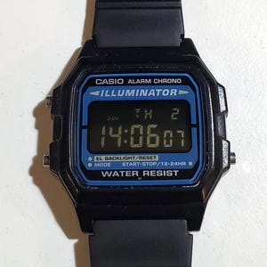 May include: Black digital Casio watch with a rectangular face and black band. The watch face displays the time, day, and date in a blue-tinted digital display. The watch has the text "CASIO ALARM CHRONO ILLUMINATOR WATER RESIST".