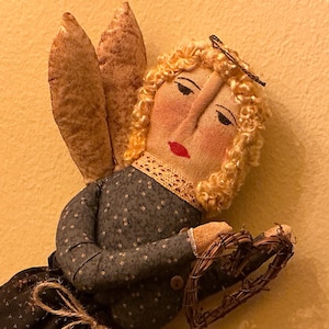 May include: A handmade angel doll with a fabric body, curly blonde hair, and brown fabric wings. The doll wears a blue dress with white polka dots and holds a woven heart. The angel has a painted face with red lips.
