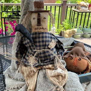 May include: A fabric scarecrow doll wearing a brown top hat, a navy and tan plaid shirt, and tan trousers, featuring a black crow on its shoulder. Beside it are two smaller round fabric pumpkin-shaped dolls with button eyes and stitched faces.