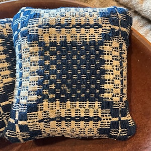 May include: Two square throw pillows featuring a woven geometric pattern in deep navy blue and natural beige. The fabric displays a textured, raised weave with a repeating grid design, creating a structured look across the surface of each pillow.