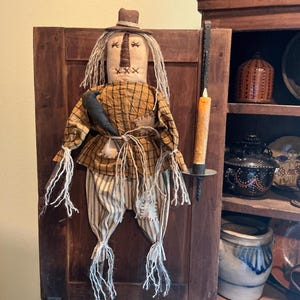 May include: A fabric scarecrow doll wearing a brown hat, a yellow and black plaid shirt, and beige striped pants. The doll features stitched facial details, frayed yarn hair, and a black crow accessory. A battery-operated candle is mounted nearby.