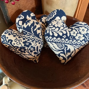 May include: A collection of three heart-shaped fabric ornaments featuring a dark navy blue and cream woven floral pattern. The hearts are filled and have a textured, tapestry-like appearance with intricate botanical designs across the surface.
