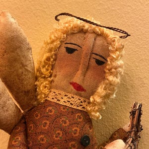 May include: A handmade angel doll with a fabric body, curly blonde hair, and painted facial features. The doll wears a brown patterned dress and has fabric wings. A halo sits atop the head. The doll is holding a bundle of twigs.