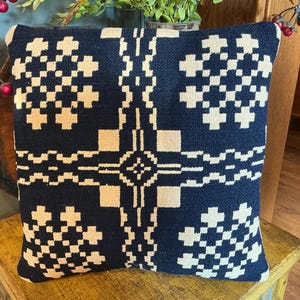 May include: A square throw pillow featuring a dark navy blue fabric base with a contrasting cream-colored woven geometric pattern. The design consists of a central cross motif surrounded by four symmetrical block-like shapes in the corners.