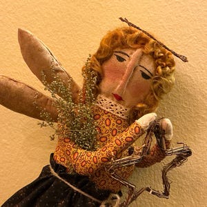 May include: A handmade angel doll with fabric wings and a star. The doll has a painted face, curly blonde hair, and wears a patterned orange and yellow dress with a black skirt. The angel holds a wire star and has a twig halo.