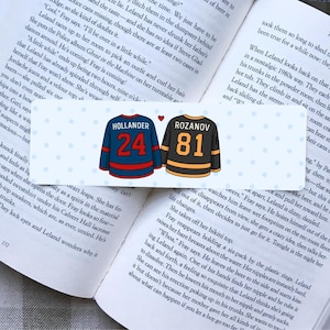 May include: Bookmark featuring two hockey jerseys, one blue with "HOLLANDER 24" and the other black with "ROZANOV 81". A small red heart sits above the jerseys. The bookmark is set against the backdrop of an open book.