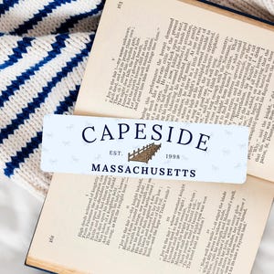 May include: A rectangular sticker with the text "CAPESIDE EST. 1998 MASSACHUSETTS" and a bridge illustration, placed on an open book with a blue and white striped sweater in the background.