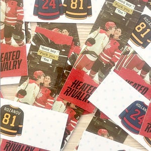 May include: A collection of hockey-themed cards and stickers. The cards feature images of hockey players and jerseys with the names "HOLLANDER" and "ROZANOV" and numbers "24" and "81". Some cards have text that reads "HEATED RIVALRY".