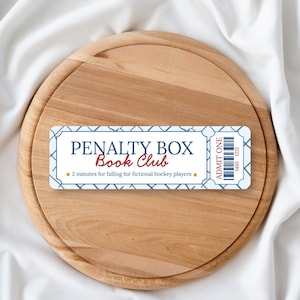 May include: A wooden cutting board with a decorative ticket that reads "PENALTY BOX Book Club". The ticket has a blue and white design with the text "2 minutes for falling for fictional hockey players" and "ADMIT ONE".