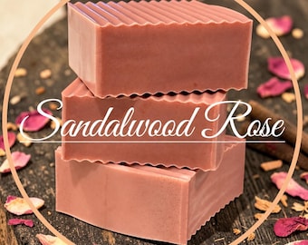 May include: Three rectangular bars of dusty rose-colored soap stacked vertically. Each bar features a textured, ridged top edge. The text Sandalwood Rose is printed in white cursive script across the center of the middle soap bar.