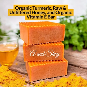 May include: Three stacked orange soap bars with textured tops, labeled "A and Shay." The image includes text: "Organic Turmeric, Raw & Unfiltered Honey, and Organic Vitamin E Bar." A glass of liquid and turmeric powder are also visible.