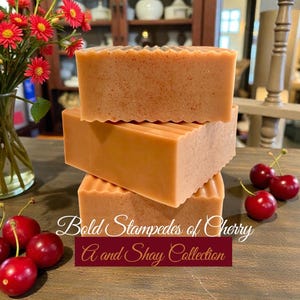 May include: Three stacked, rectangular bars of soap with a textured surface and a light brown color. The image also features fresh red cherries and a vase of red flowers. Text on the image reads "Bold Stampedes of Cherry A and Shay Collection."