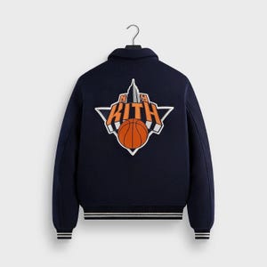 May include: A navy blue varsity jacket featuring a large embroidered graphic on the back. The design includes the text KITH in orange letters, a grey skyscraper illustration, and an orange basketball, all set against a white triangular background.