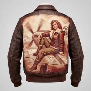 Pode incluir: Uma jaqueta bomber de couro marrom com um gráfico grande e detalhado nas costas de uma pessoa com cabelo ondulado sentada na asa de um avião de hélice vintage. A jaqueta tem gola clássica, punhos e bainha canelados em tom marrom.
