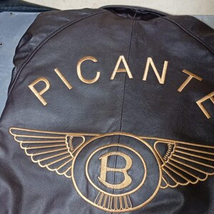 Puede incluir: Chaqueta de cuero negra con texto y emblema bordados en dorado. El texto dice "PICANTE" encima de un emblema circular con alas y la letra "B" en su interior, y "BENTLEY" debajo.