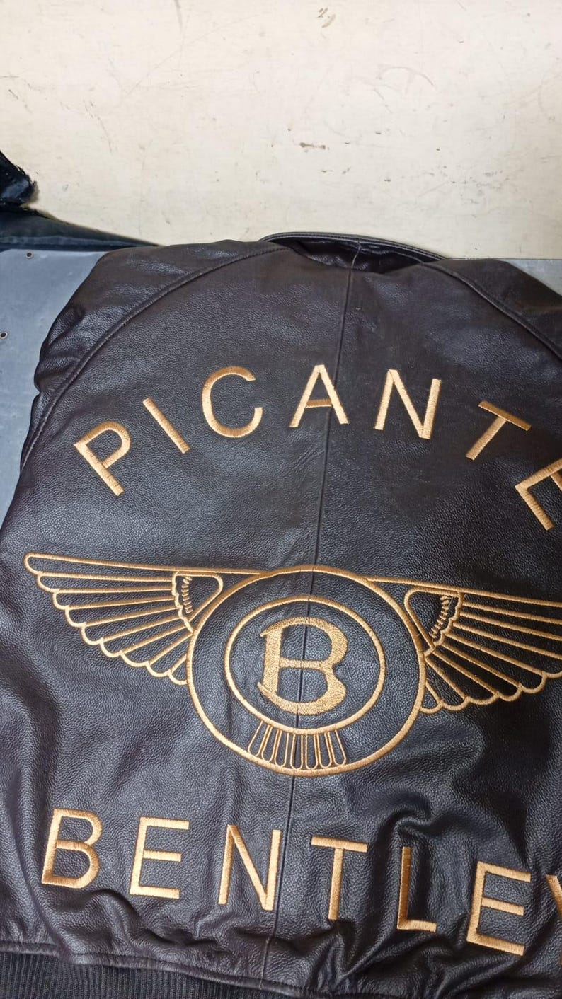Puede incluir: Chaqueta de cuero negra con texto y emblema bordados en dorado. El texto dice "PICANTE" encima de un emblema circular con alas y la letra "B" en su interior, y "BENTLEY" debajo.