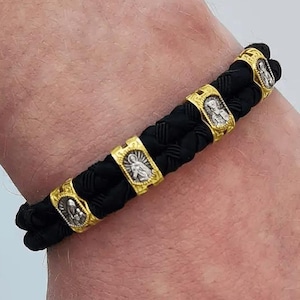 May include: Black braided bracelet with gold-colored accents. The bracelet features several gold-toned rectangular beads with religious iconography. The bracelet is worn on a wrist, showcasing its design and craftsmanship. The bracelet is a religious accessory.