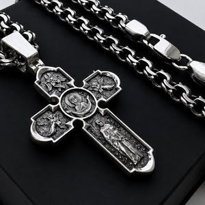 May include: A silver-colored Orthodox cross pendant with intricate religious iconography, suspended from a matching chain. The cross features detailed engravings and is displayed on a black surface, showcasing the jewelry's design.