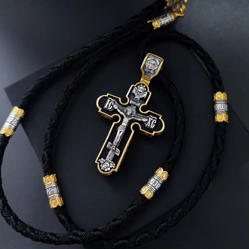 May include: A gold and silver cross pendant with a black braided cord. The cross features a detailed depiction of Jesus Christ and religious text. The cord has gold and silver accents. The pendant is likely made of precious metals.