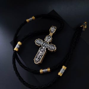 May include: A silver and gold-toned Orthodox cross pendant with intricate detailing and the inscription "Спаси и Сохрани". The cross is suspended on a black braided cord with gold and silver accents. The pendant features religious iconography.