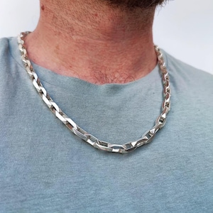 May include: A silver-colored chain necklace with a chunky, rectangular link design. The necklace is worn around the neck and rests on a light blue t-shirt. The chain has a polished finish, reflecting light.