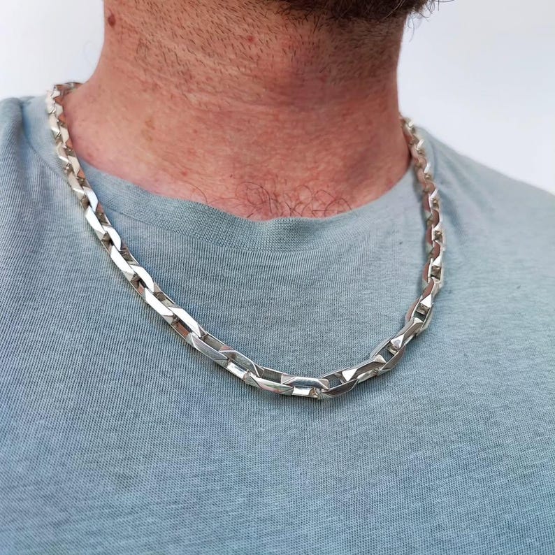 May include: A silver-colored chain necklace with a chunky, rectangular link design. The necklace is worn around the neck and rests on a light blue t-shirt. The chain has a polished finish, reflecting light.